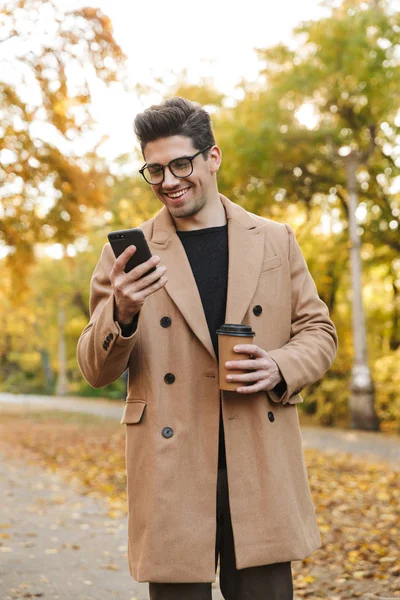 Image of happy young man wearing coat typing on smartphone and smiling in autumn park — Stock Photo, Image