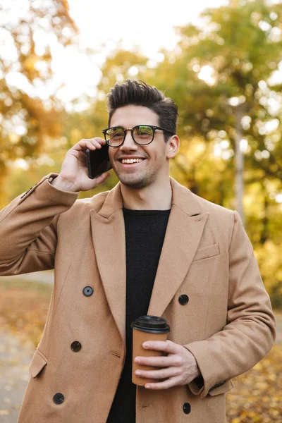 Образ щасливого молодого чоловіка в пальто розмовляє на смартфоні і посміхається в осінньому парку — стокове фото