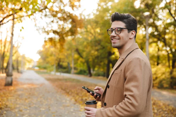 Retrato de homem morena elegante vestindo casaco digitando no smartphone e sorrindo no parque de outono — Fotografia de Stock