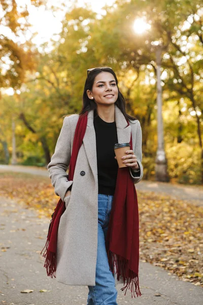 Retrato de una joven morena con abrigo bebiendo café para llevar y sonriendo a la cámara en el parque de otoño —  Fotos de Stock