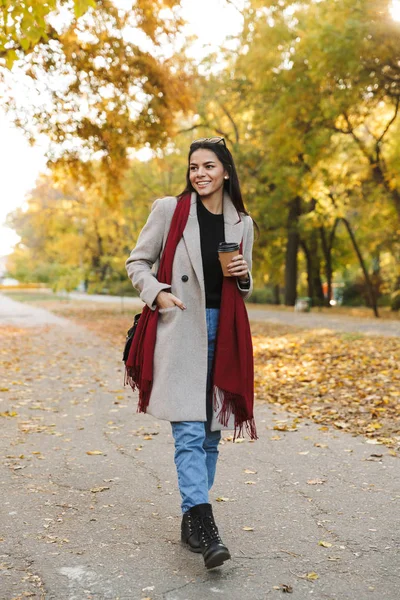 Portrait of stylish brunette woman wearing coat drinking takeaway coffee and smiling in autumn park — Stock Photo, Image
