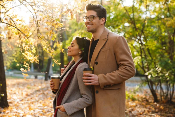 Retrato de pareja romántica tomando café para llevar y abrazándose mientras camina en el parque de otoño —  Fotos de Stock