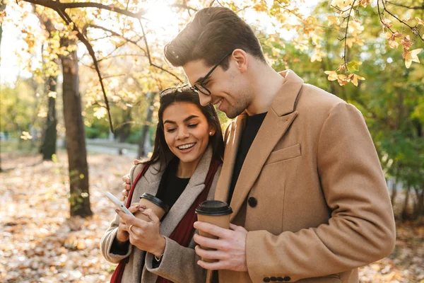 Porträt eines stilvollen Paares, das im Herbstpark Kaffee zum Mitnehmen trinkt und beim Spazierengehen aufs Smartphone schaut — Stockfoto