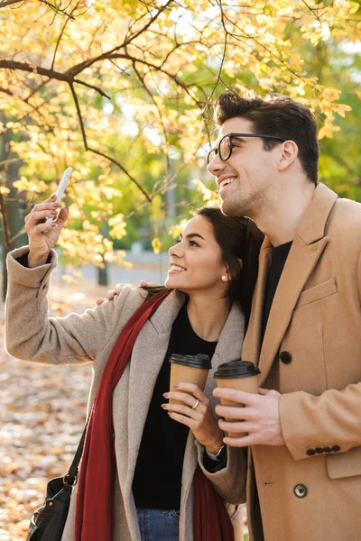 Retrato de una hermosa pareja bebiendo café para llevar y tomando una foto selfie en el teléfono celular mientras camina en el parque de otoño —  Fotos de Stock