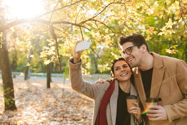Retrato de una pareja sonriente tomando café para llevar y tomando una foto selfie en el teléfono celular mientras camina en el parque de otoño —  Fotos de Stock