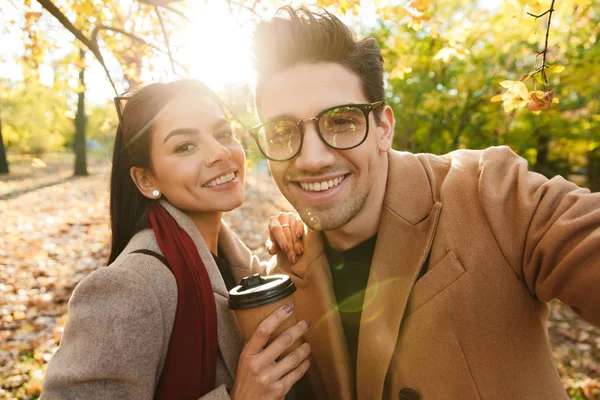 Retrato de pareja feliz tomando café para llevar y sonriendo a la cámara mientras camina en el parque de otoño —  Fotos de Stock