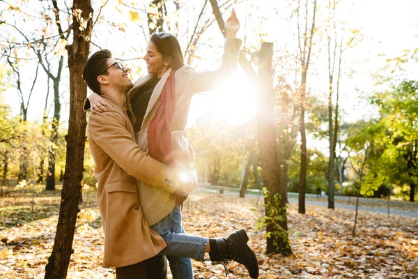 Imagen de un hombre guapo llevando a una mujer en las manos y sonriendo en el parque de otoño — Foto de Stock