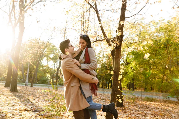 Imagen de un hombre joven llevando a una mujer en las manos y sonriendo en el parque de otoño — Foto de Stock