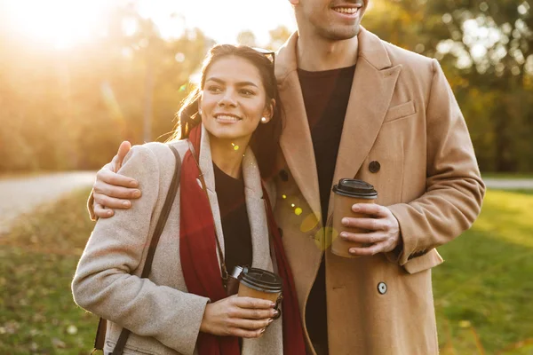 Retrato de pareja atractiva bebiendo café para llevar de tazas de papel mientras camina en el parque de otoño —  Fotos de Stock