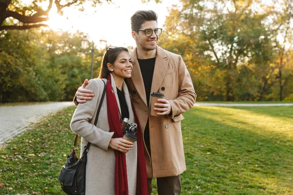 Retrato de una alegre pareja tomando café para llevar de las tazas de papel mientras camina en el parque de otoño —  Fotos de Stock