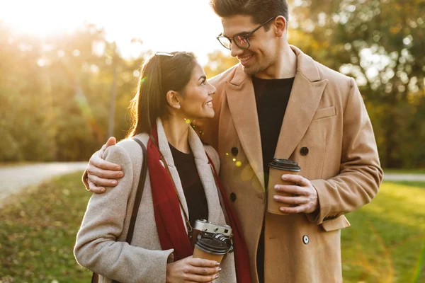 Portrait of european couple drinking takeaway coffee and looking at each other while walking in autumn park — Stock Photo, Image