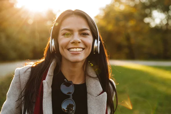 Retrato de una joven morena con abrigo escuchando música con auriculares y sonriendo en el parque de otoño —  Fotos de Stock