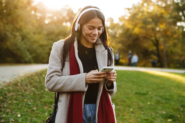 ヘッドフォンで音楽を聴いたり、公園で携帯電話を使用してコートを着て幸せな素敵な女性の肖像画 — ストック写真