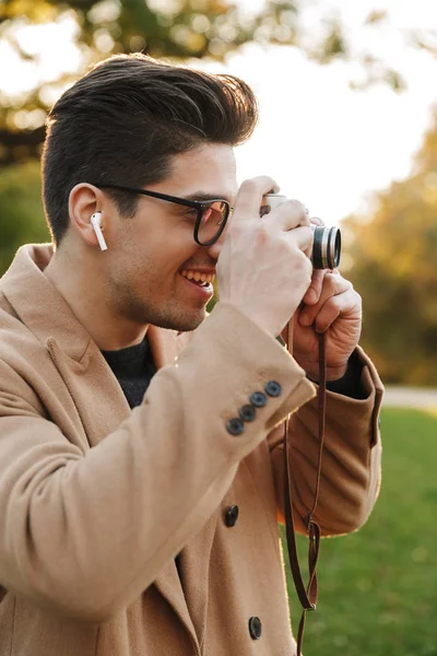 Foto en el perfil del hombre alegre escuchando música con auricular y tomando fotos en la cámara retro en el parque de otoño — Foto de Stock