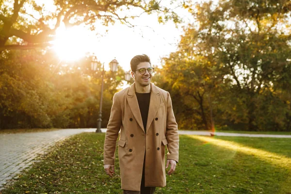 Foto de un buen hombre caucásico escuchando música con auricular y sonriendo mientras camina en el parque de otoño — Foto de Stock