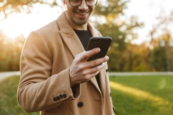 Photo of smiling caucasian man listening to music with earpods and typing on smartphone in autumn park — Stock Photo, Image