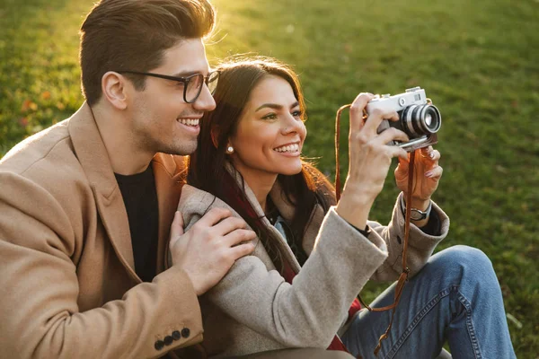 Retrato de pareja sonriente tomando fotos en cámara retro mientras está sentada en el césped en el parque —  Fotos de Stock