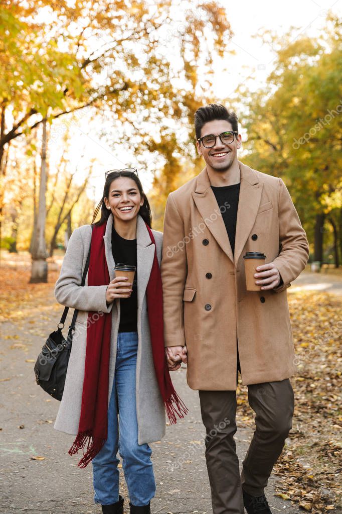 Portrait of casual couple drinking takeaway coffee and smiling at camera while walking in autumn park