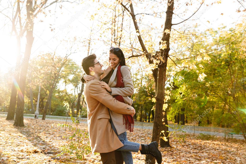Image of young man carrying woman in hands and smiling in autumn park