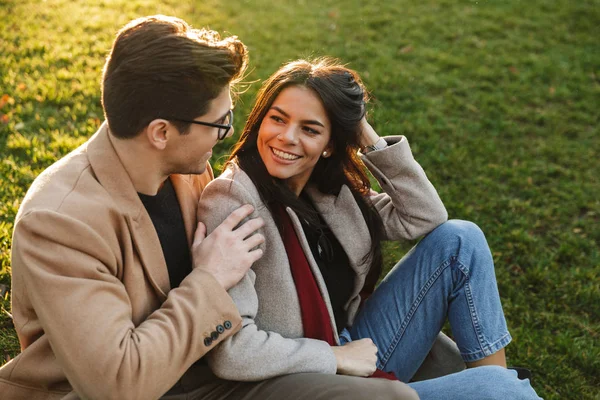 Imagen de pareja caucásica morena sonriendo y abrazándose mientras está sentada en la hierba en el parque —  Fotos de Stock