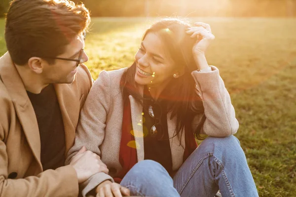 Imagen de una linda pareja caucásica abrazándose y mirándose mientras están sentados en el césped en el parque —  Fotos de Stock