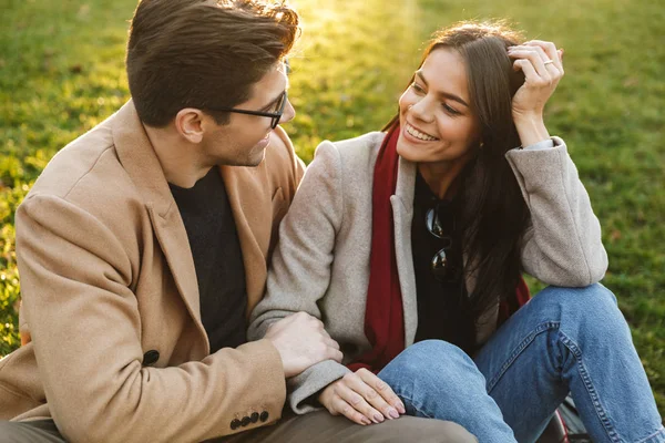 Bild von niedlichen europäischen Paar umarmen und einander anschauen, während sie auf Gras im Park sitzen — Stockfoto