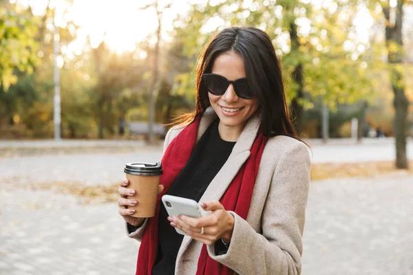 Retrato de mulher morena sorridente usando óculos escuros bebendo café e digitando no celular no parque de outono — Fotografia de Stock