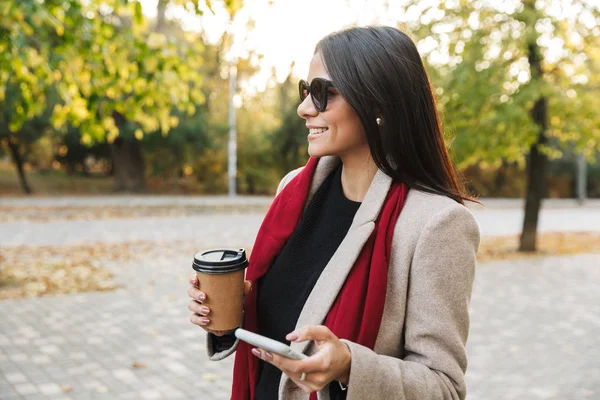 Retrato de una joven sonriente con gafas de sol bebiendo café para llevar y escribiendo en el teléfono celular en el parque de otoño — Foto de Stock