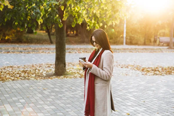 Portret van glimlachend stijlvolle vrouw dragen zonnebril drinken afhaalkoffie en typen op mobiele telefoon in herfst Park — Stockfoto