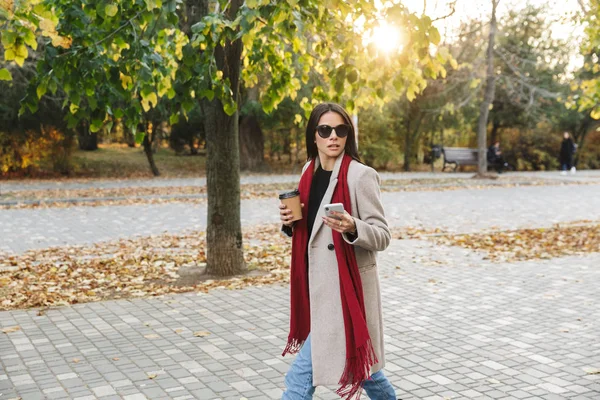 Portrait of thinking stylish woman wearing sunglasses drinking takeaway coffee and typing on cellphone n autumn park — Stock Photo, Image