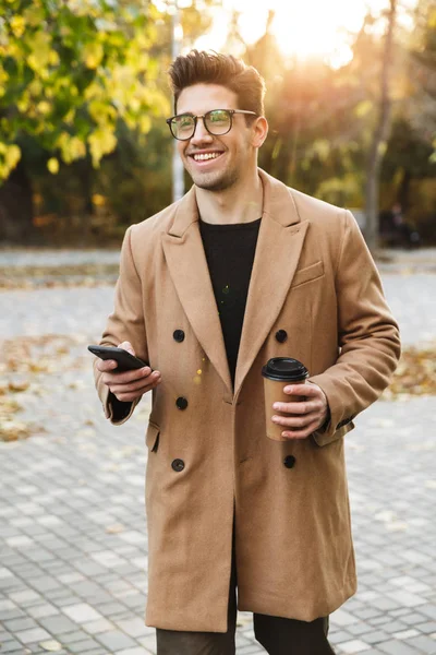 Image of handsome smiling man wearing coat using cellphone and drinking takeaway coffee in autumn park — Stock Photo, Image