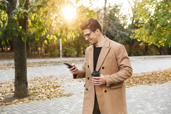 Зображення гарного випадкового чоловіка в пальто з використанням мобільного телефону та виймання кави в осінньому парку — стокове фото