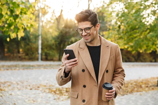 Image of handsome smiling man wearing coat typing cellphone and drinking takeaway coffee in autumn park — Stock Photo, Image