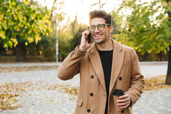 Образ молодого усміхненого чоловіка в пальто, що розмовляє на мобільному телефоні та виймає каву в осінньому парку — стокове фото