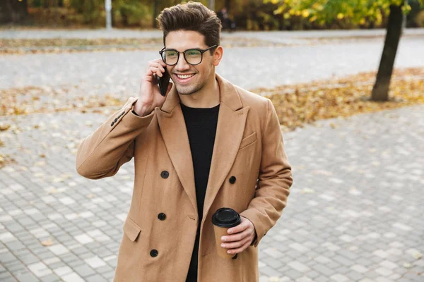 Obraz stylowy uśmiechnięty człowiek nosi płaszcz rozmawia na telefon komórkowy i picie taimage stylowy uśmiechnięty człowiek ubrany w płaszcz kawy na wynos w jesiennym parku — Zdjęcie stockowe
