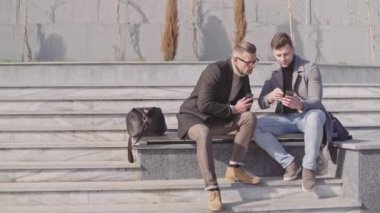 Gündelik iki yakışıklı yetişkin erkek dışında bir bankta otururken akıllı telefonlar kullanıyor