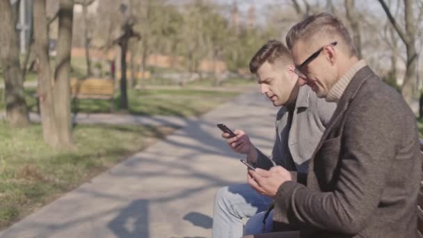 Двоє Красивих Дорослих Чоловіків Смартфонами Розмовляють Сміються Сидячи Парку — стокове відео