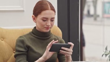 Kafede oturan akıllı telefon oyunu oynayan güzel genç kızıl saçlı kadın