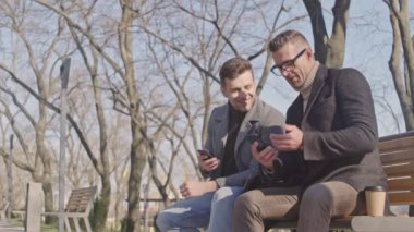 Parkta otururken akıllı telefon kullanarak konuşan iki gülümseyen yetişkin erkek