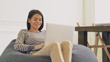 Bir beanbag sandalyede oturan dizüstü bilgisayar kullanarak Gülümseyen genç kadın