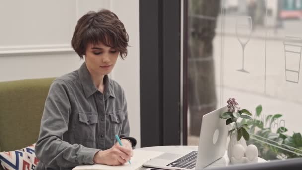 カフェに座ってメモ帳に何かを指摘する魅力的な若い女性 — ストック動画