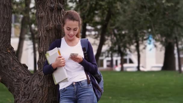 Щаслива Брюнетка Рюкзаком Книгами Використовуючи Свій Смартфон Стоячи Біля Дерева — стокове відео