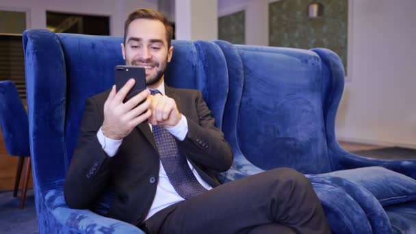 カフェでアームチェアに座りながらスマートフォンを使ってあごひげを生やしたビジネスマンを喜ばせる — ストック動画