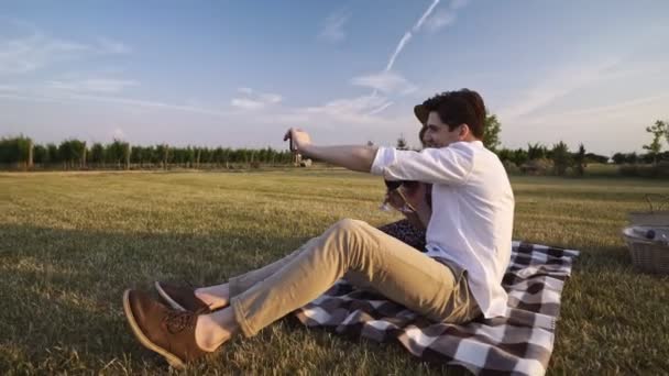 お互いに話し合うワインを飲んでフィールドで屋外に座っている若い幸せな愛するカップルは 携帯電話で自分撮りを取ります — ストック動画