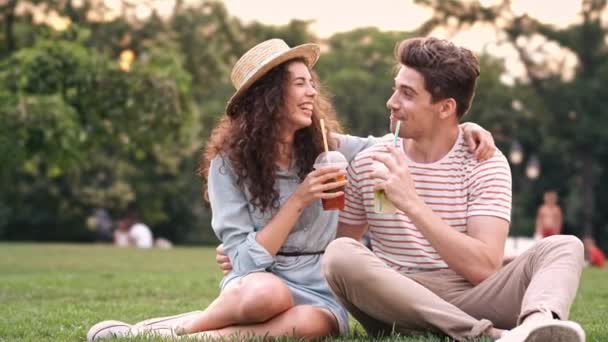幸せなロマンチックなカップルは 草の上に座って お互いにジュースカクテルを飲んで話しています — ストック動画