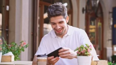 Sokakta açık havada kafede oturan genç mutlu yakışıklı adam kahve içerek cep telefonu ile oyun oynamak