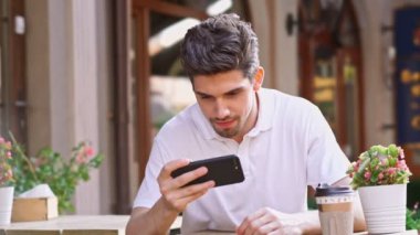 Genç mutlu yakışıklı adam sokakta kafede oturan cep telefonu içme kahve kullanarak kazanan jest yapmak