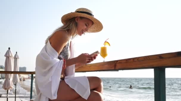 华丽的金发女郎 年代在夏天草帽有一个休息在海滩上喝鸡尾酒用手机 — 图库视频影像