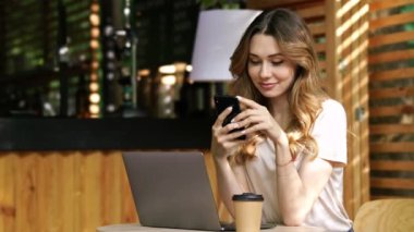 Kafe açık havada akıllı telefon kullanırken dizüstü bilgisayar ile masanın yanında oturan rahat giysiler içinde Gülümseyen esmer kadın