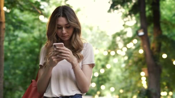 カジュアルな服を着た笑顔のブルネット女性が屋外に立っている間 スマートフォンにメッセージを書き込む — ストック動画